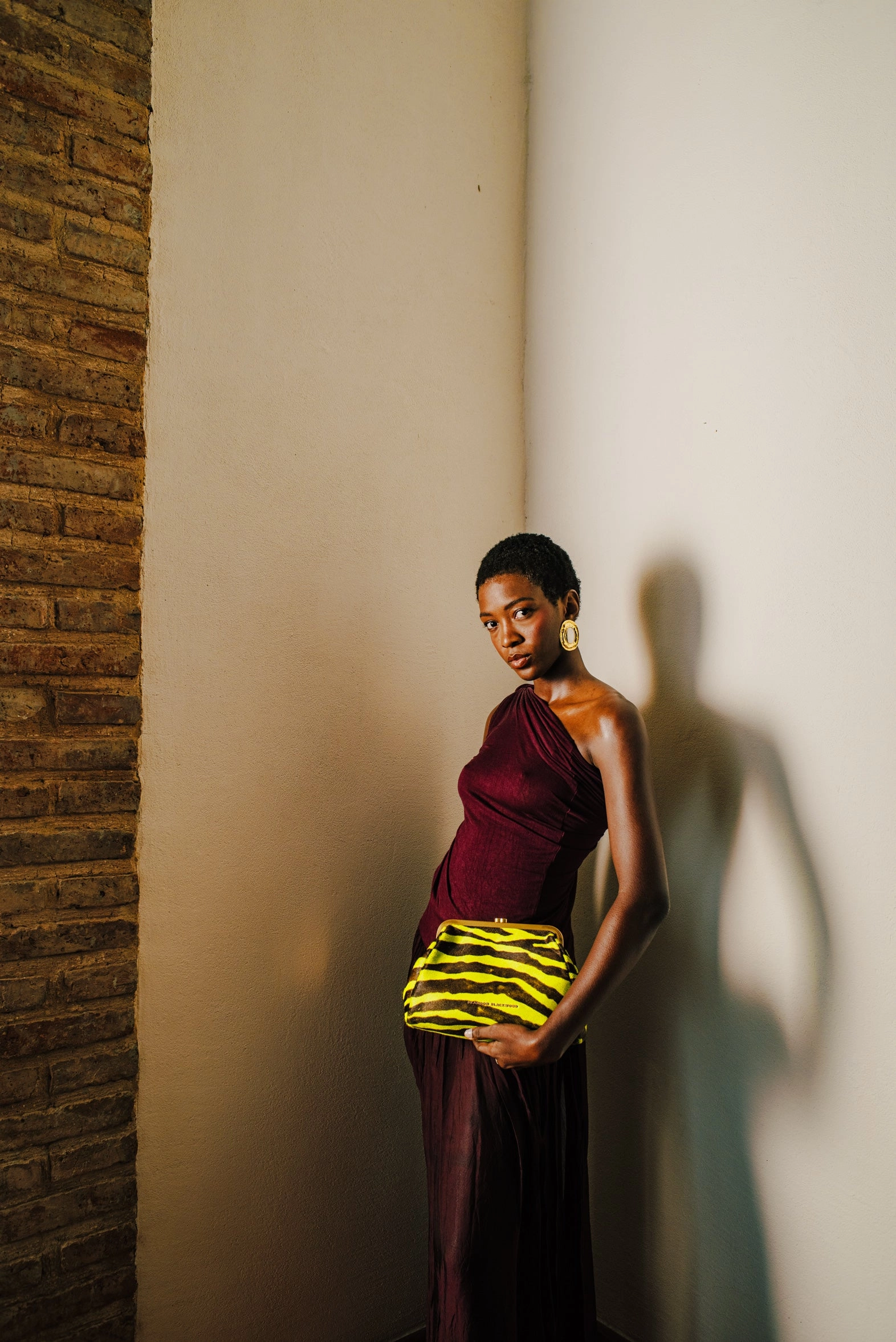A woman in a burgundy one-shoulder dress stands by a brick wall holding a yellow zebra clutch