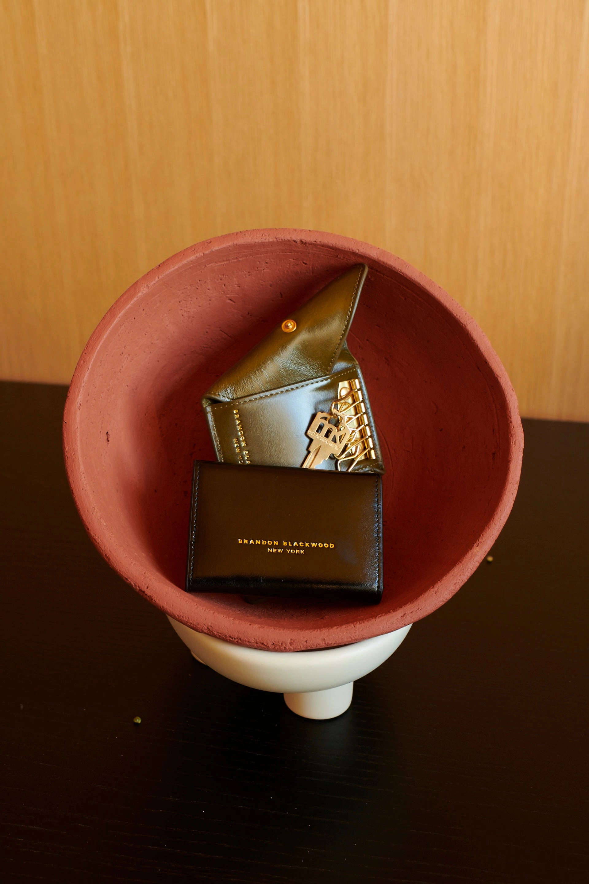 A black Brandon Blackwood New York wallet and an olive key pouch with a gold key charm in a terracotta bowl