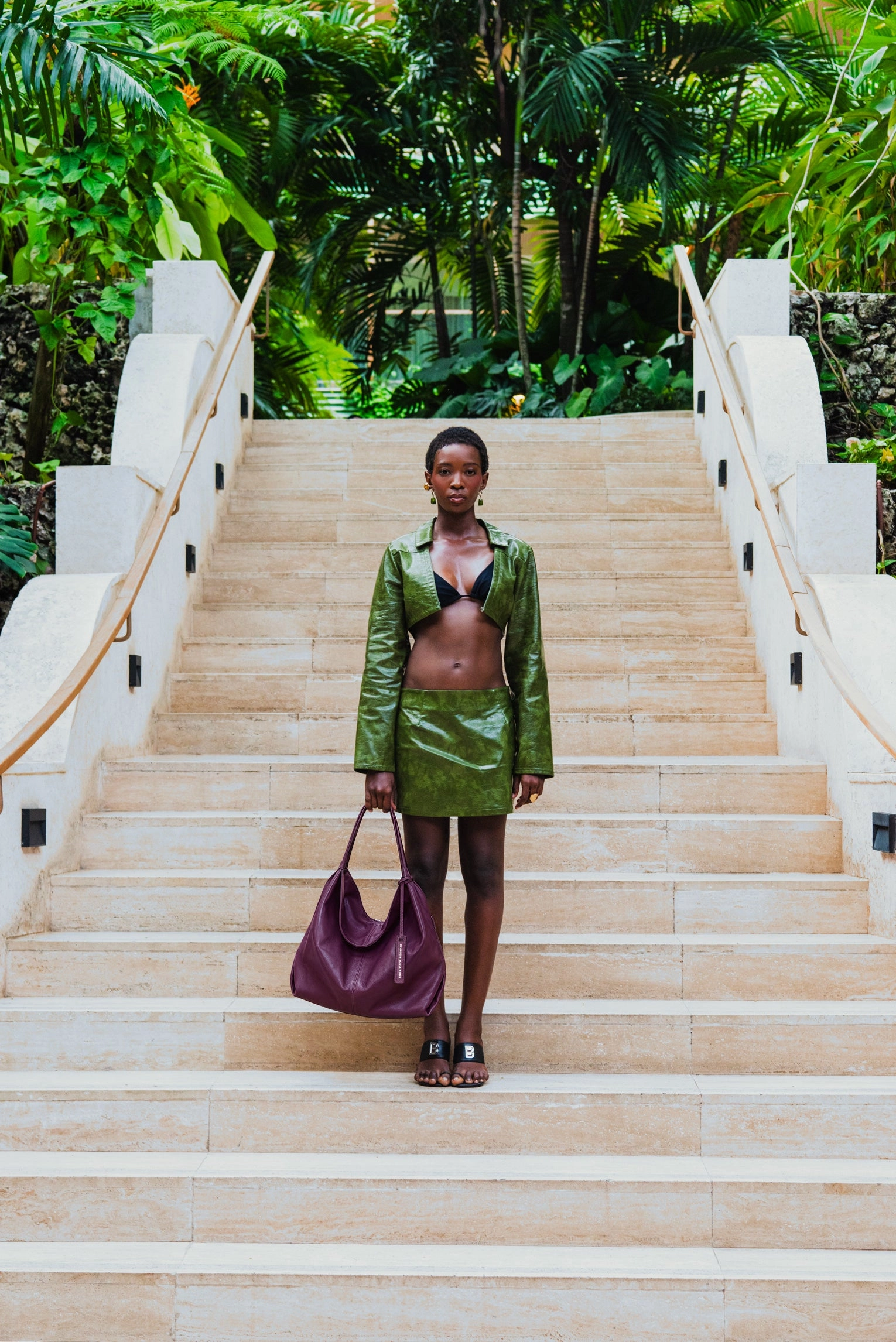 A woman in a green leather set stands on a wide stone staircase holding an eggplant leather shoulder bag