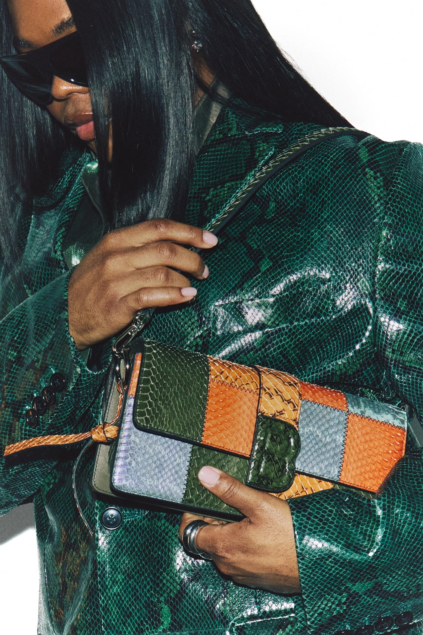A hand with silver rings holds a patchwork snakeskin bag against a dark green croc-embossed jacket