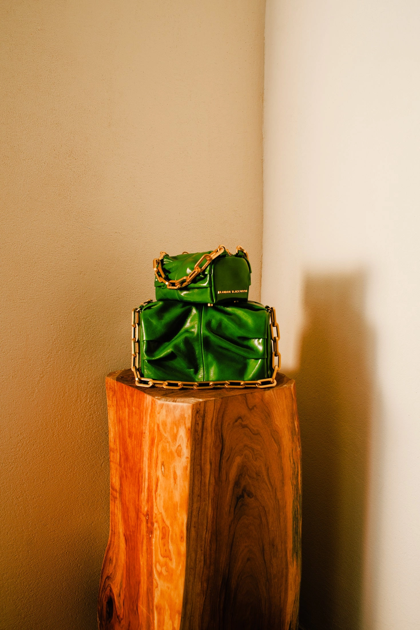Two green oiled leather bags with brass chains stacked on a carved wooden plinth