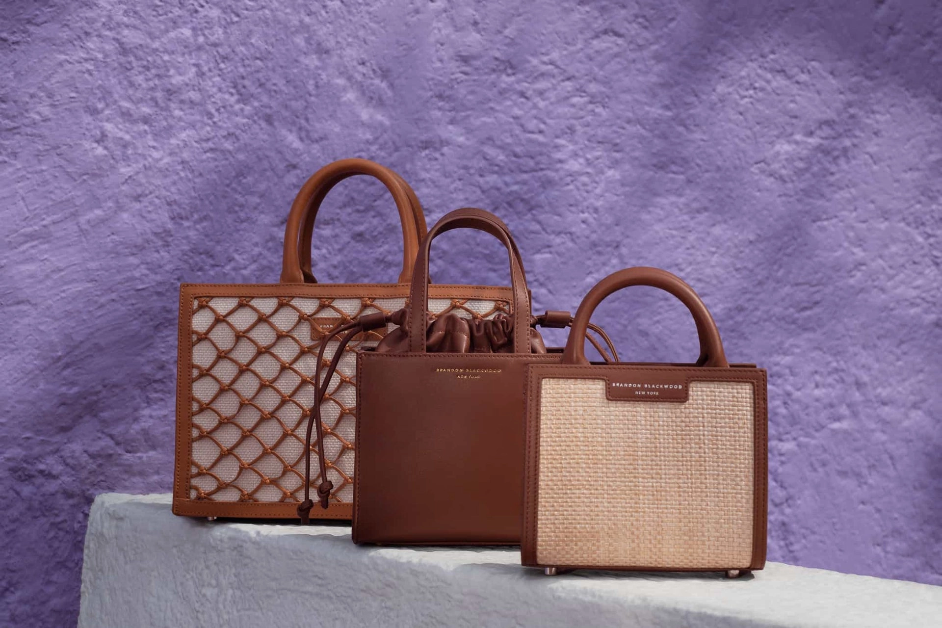 Three structured tote bags in brown leather and woven panels on a draped plinth against a violet wall