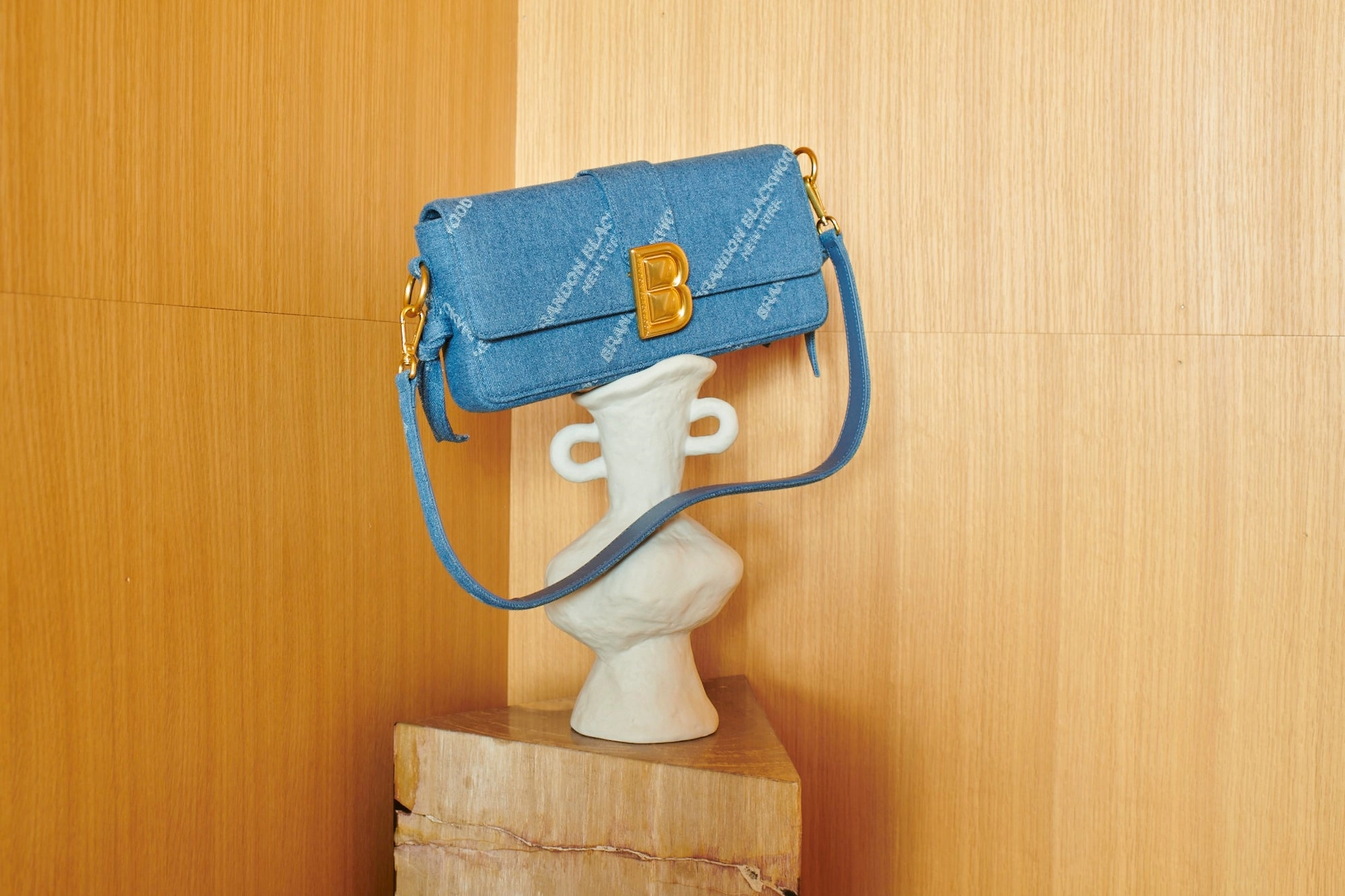 A blue quilted shoulder bag with a gold B clasp hanging from a white ceramic vase against a wooden wall